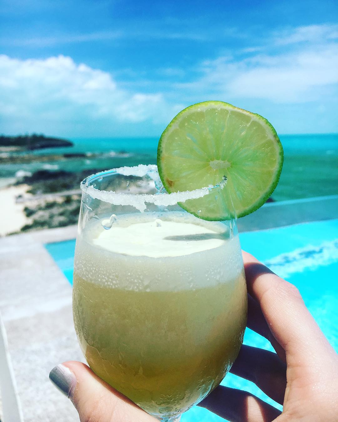 Woman holding a margarita over the pool, with the ocean as a backdrop. 