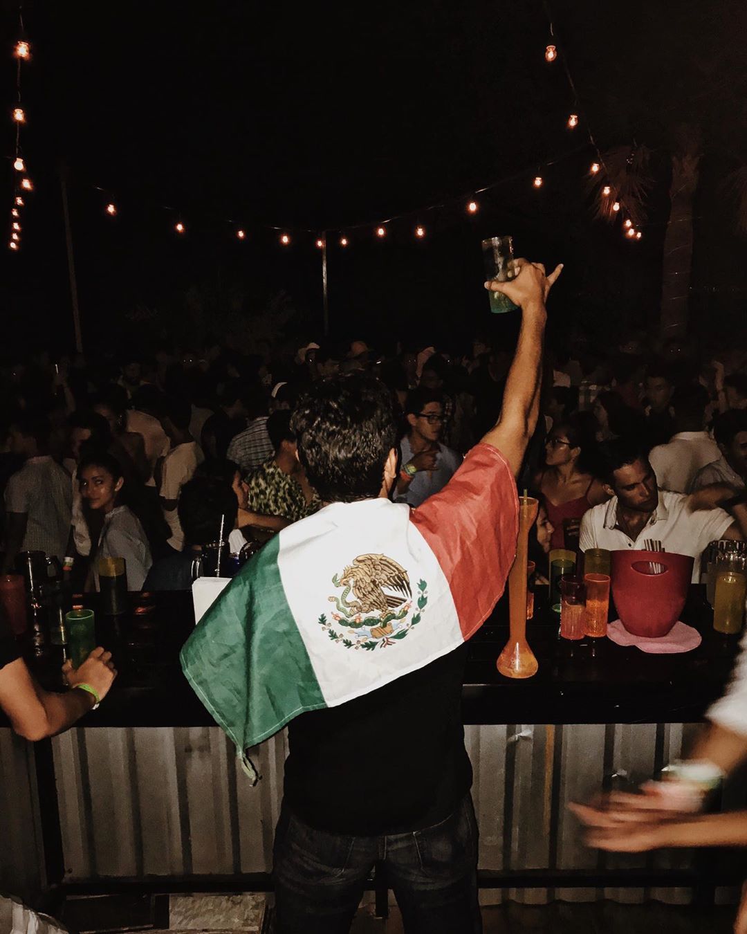 Man raising glass in front of crowd with Mexican flag on his back.